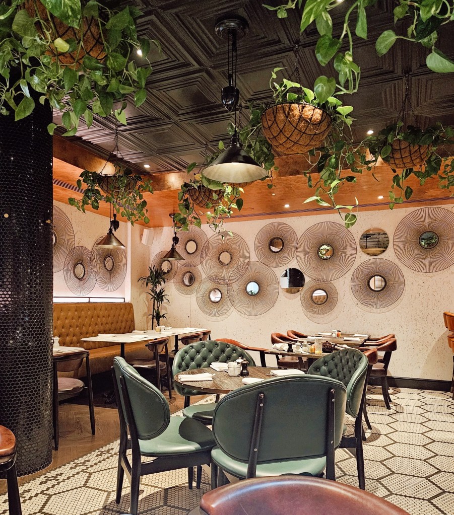 Interior of a stylish restaurant featuring green plants, elegant seating, and decorative wall mirrors in a relaxed atmosphere.