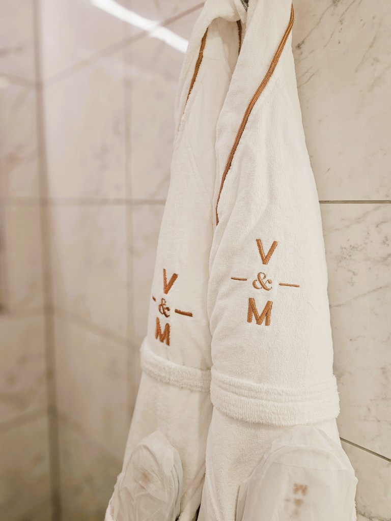 Two white bathrobes with embroidered initials 'V & M' hanging on a hook against a tiled bathroom wall.