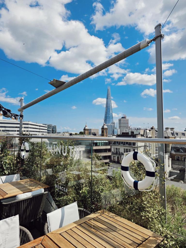 View from the Mercer Roof Terrace at Vintry & Mercer hotel, showcasing outdoor seating with wooden tables, greenery, and the London skyline including The Shard under a blue sky with fluffy clouds.