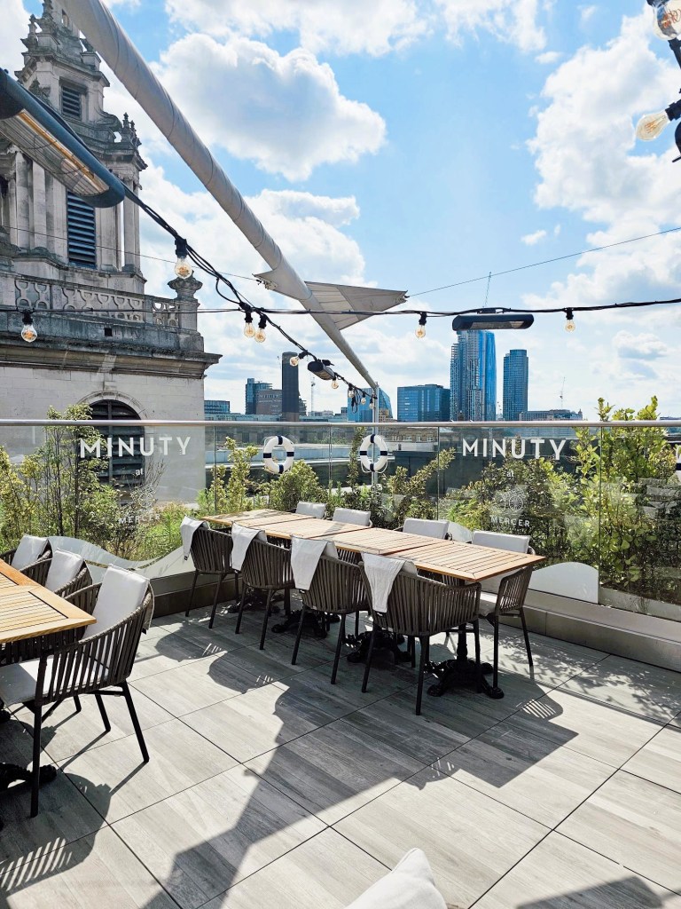 A rooftop terrace at Vintry & Mercer hotel in London, featuring wooden dining tables and chairs, surrounded by greenery and a view of the city skyline under a blue sky with scattered clouds.
