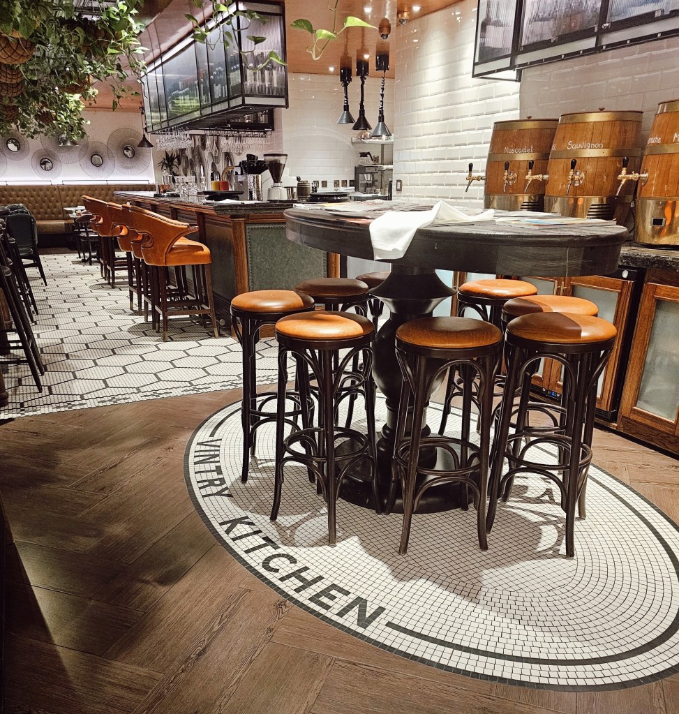 Interior view of Vintry Kitchen at Vintry & Mercer Hotel, showcasing a round table surrounded by black and brown bar stools with a stylish bar in the background.