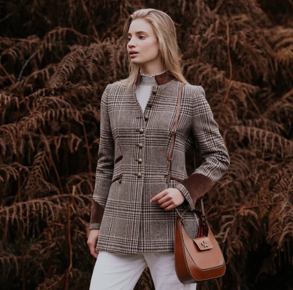 A woman in a checked patterned jacket stands against a backdrop of ferns, holding a brown handbag, exuding a sense of elegance and style.