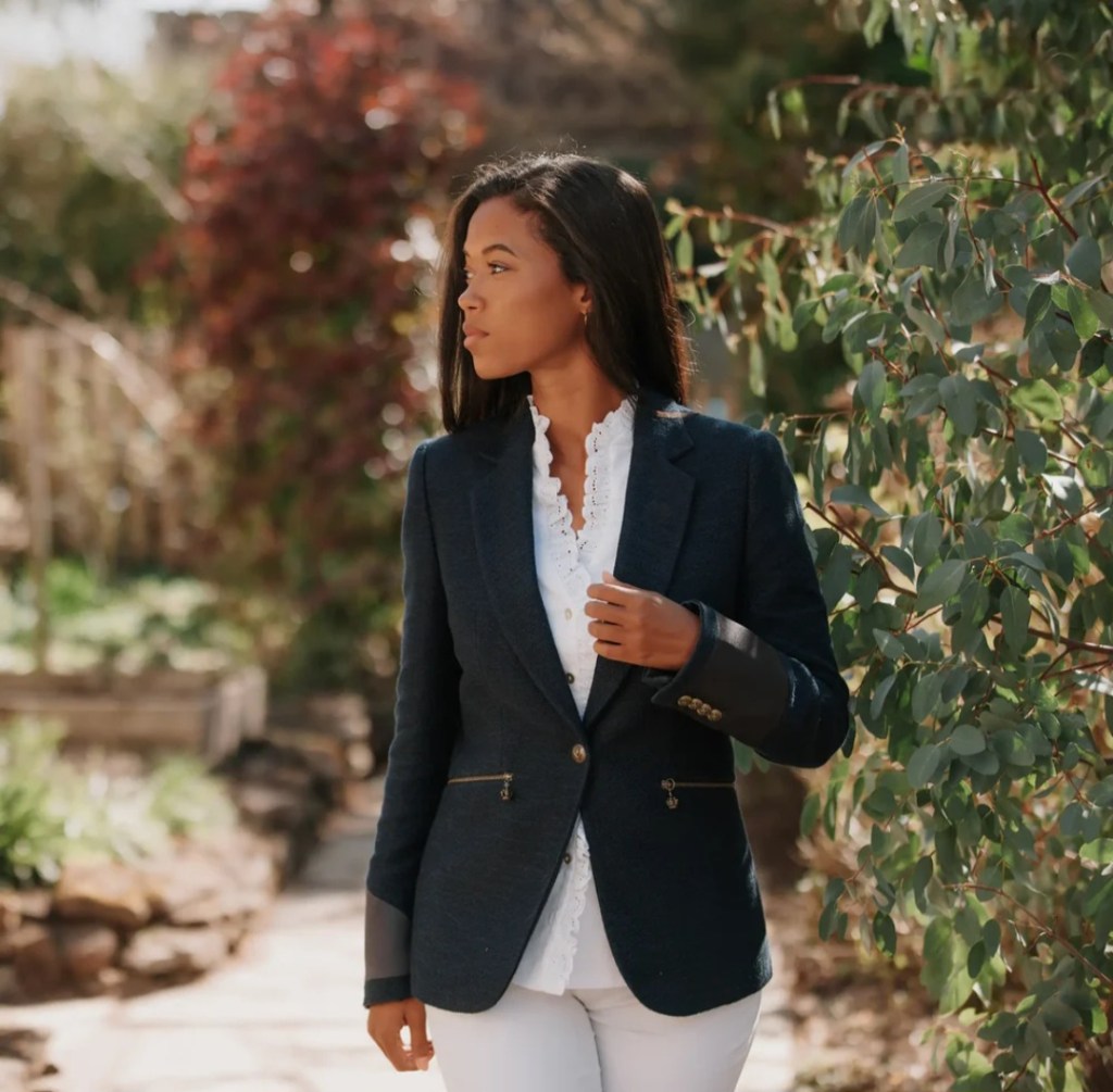 A woman wearing a stylish navy blazer over a white ruffled shirt, walking in a garden setting surrounded by greenery.