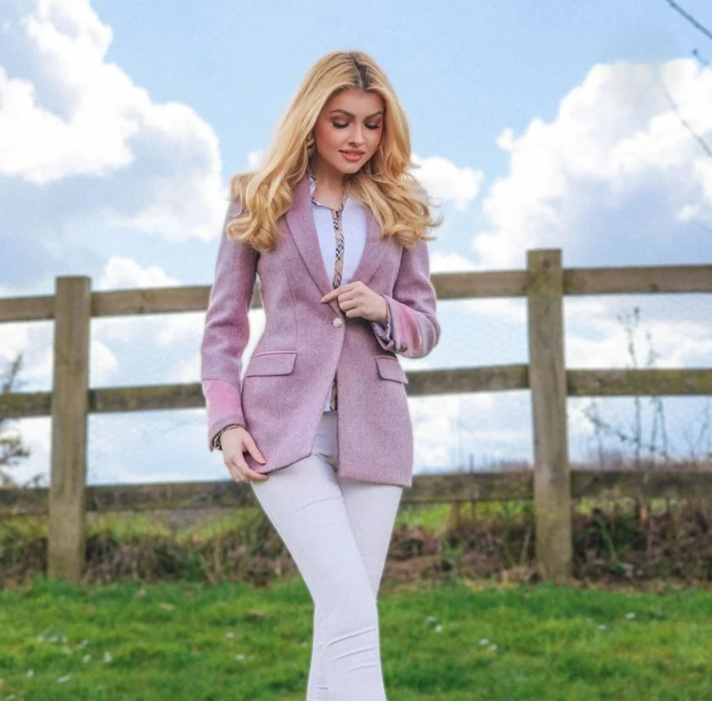 A woman wearing a pink blazer and white pants stands in a grassy field, with a wooden fence and cloudy sky in the background.