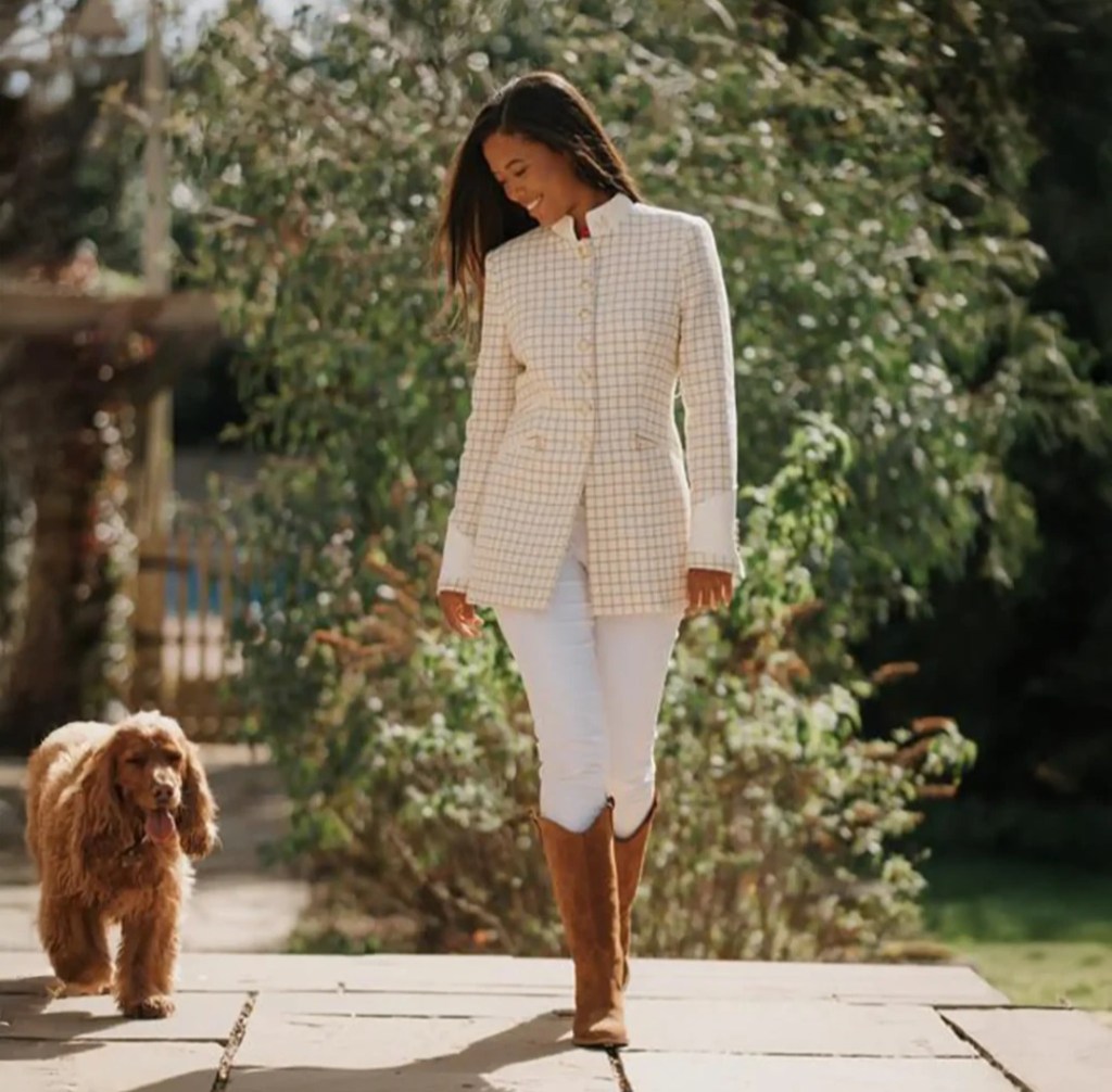 A woman wearing a stylish checked blazer, white trousers, and knee-high brown boots walks alongside a golden cocker spaniel in a lush outdoor setting.