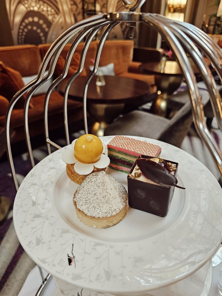 A close-up view of a tiered plate showcasing an assortment of elegant pastries, including a lemon dessert topped with a yellow sphere, a delicate flower-shaped dessert, a meringue pie, and a stylish chocolate confection, set against a luxurious lounge backdrop.