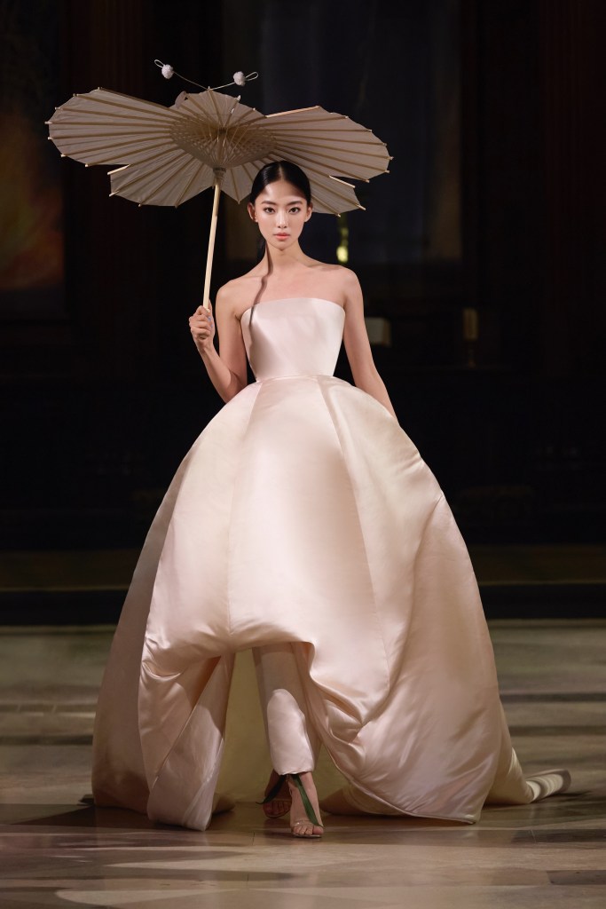 A model walks the runway at London Fashion Week, wearing a sculptural ballgown with a parasol, showcasing the elegance of Tran Hung's SS2026 collection.
