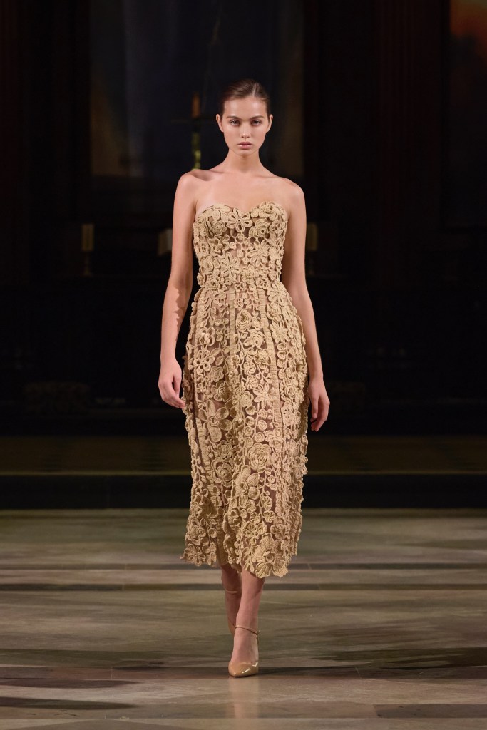 Model walking the runway in a tan, sculptural gown adorned with intricate floral embroidery at London Fashion Week.