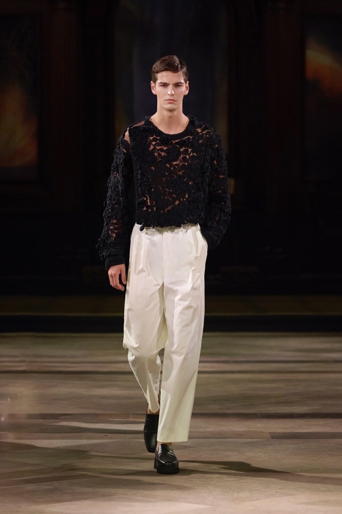 A male model walks the runway wearing a black, intricately detailed top and white trousers, showcasing modern fashion design.