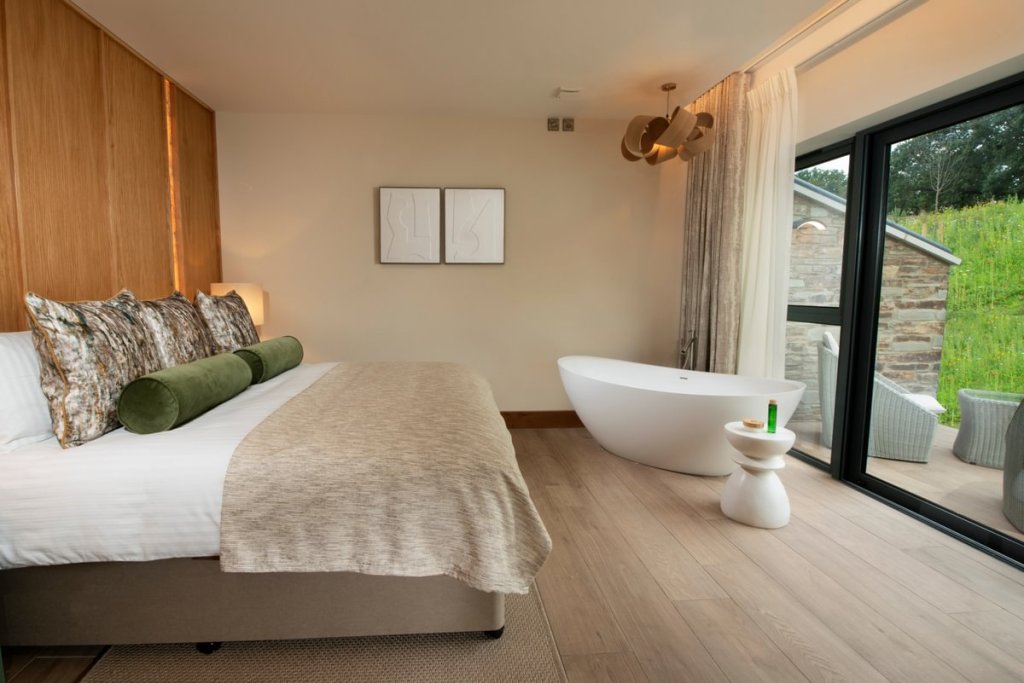 A modern Wellness Room at Boringdon Hall featuring a king-sized bed with decorative pillows, a freestanding bathtub, and large glass windows overlooking greenery.