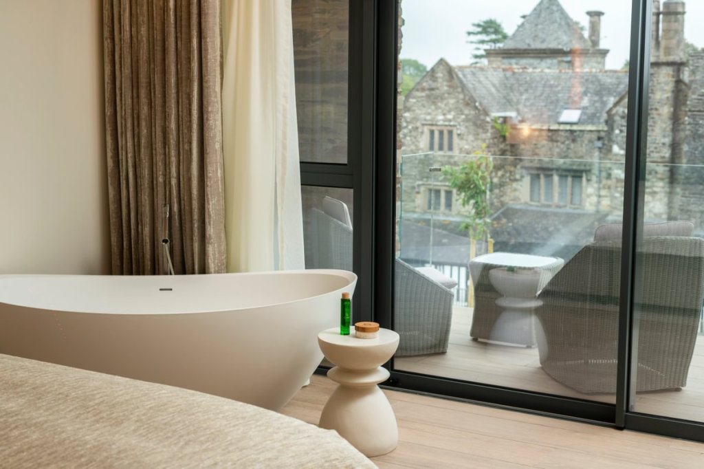 A modern wellness room at Boringdon Hall featuring a freestanding bathtub, neutral decor, and large glass windows showcasing a verdant outdoor view.