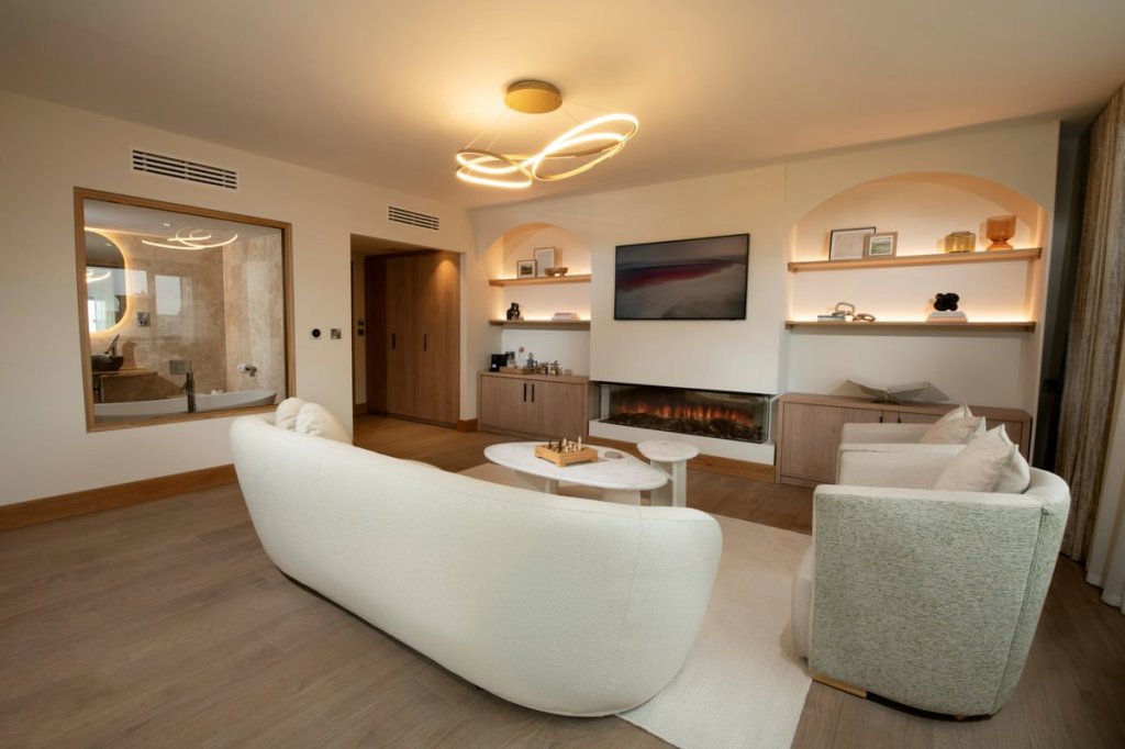 A luxurious and contemporary wellness room at Boringdon Hall, featuring a comfortable white sofa, stylish armchairs, a modern coffee table, and soft lighting, with a view of a spacious bathroom through a large mirror.