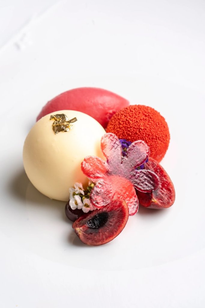 A beautifully arranged dessert featuring a white orb adorned with gold, surrounded by red and pink spherical treats and fresh fruit, including cherries and edible flowers, presented on a white plate.