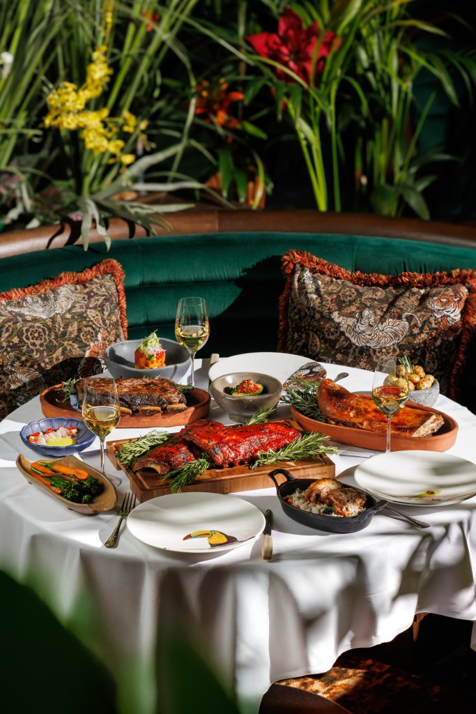A beautifully presented table featuring an array of delicious dishes from the Amazónico Asado, including grilled meats, colorful sides, and elegant glassware, set against a backdrop of lush greenery and decorative pillows.