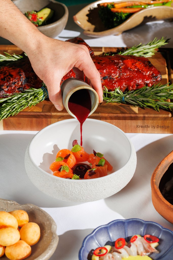 A hand pouring a rich sauce over a dish featuring orange salmon cubes, garnished with mint and black currants, surrounded by side dishes including yellow cheese balls and an array of vibrant vegetables.
