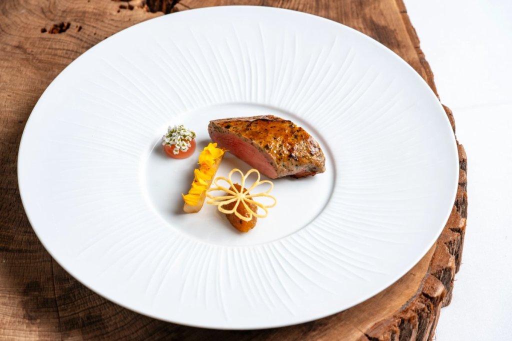 A gourmet dish featuring a succulent piece of meat paired with vibrant garnishes, elegantly presented on a decorative white plate atop a wooden surface.