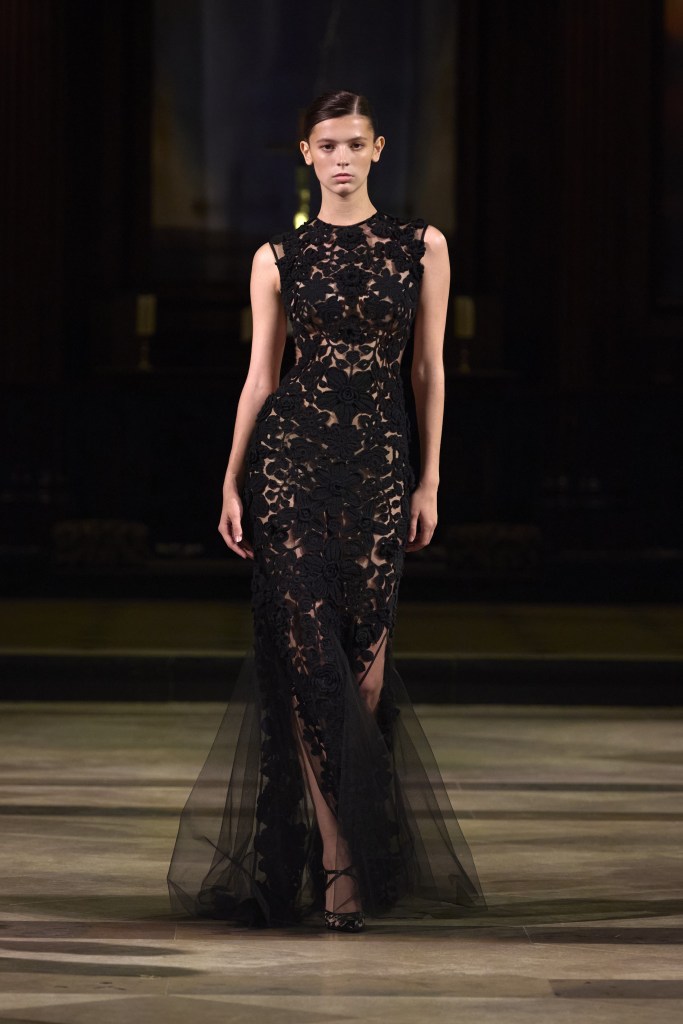 Model walking the runway wearing an intricately designed black gown with floral appliqués and sheer fabric at London Fashion Week.