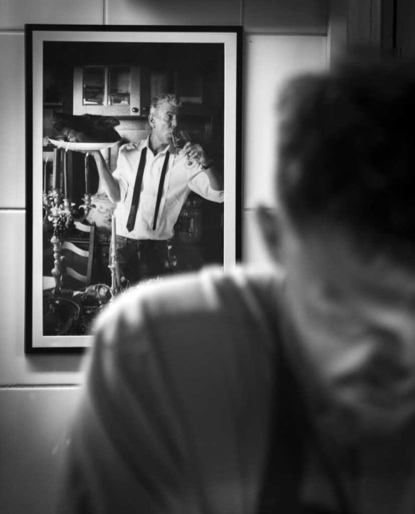 A black-and-white photo of a man in a white shirt and black tie, holding a glass while standing next to a cat on a plate. The image is framed and positioned on the wall, with a blurred figure of another person in the foreground.