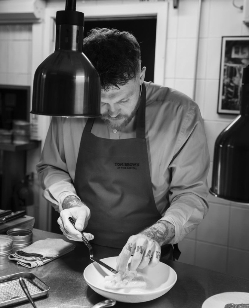 A chef, Tom Brown, is focused on plating seafood in a kitchen setting, wearing an apron with his name and restaurant. The image captures the intricate process of dish preparation.