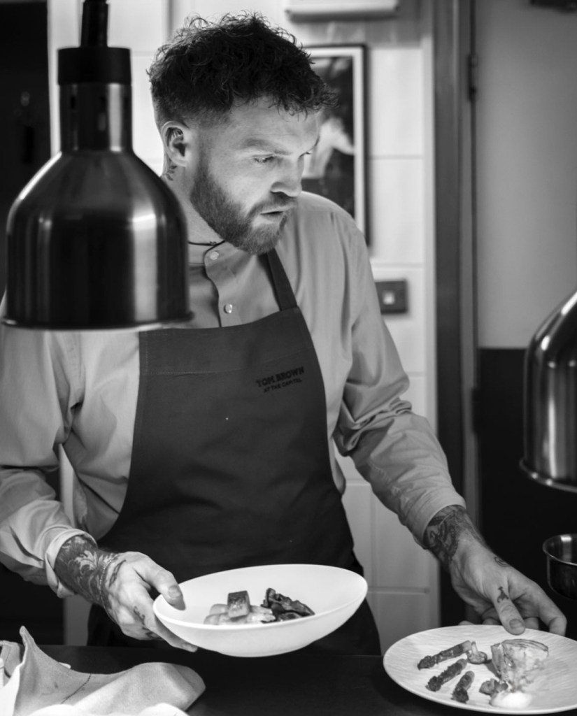 A chef carefully plating a dish in a professional kitchen, wearing a branded apron and focused on presentation.
