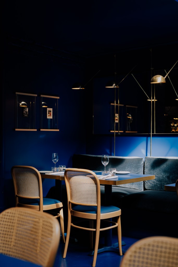 Interior view of Il Bistrotto London featuring a blue-walled dining area with a wooden table set for two, surrounded by cane-backed chairs and illuminated by stylish pendant lights.