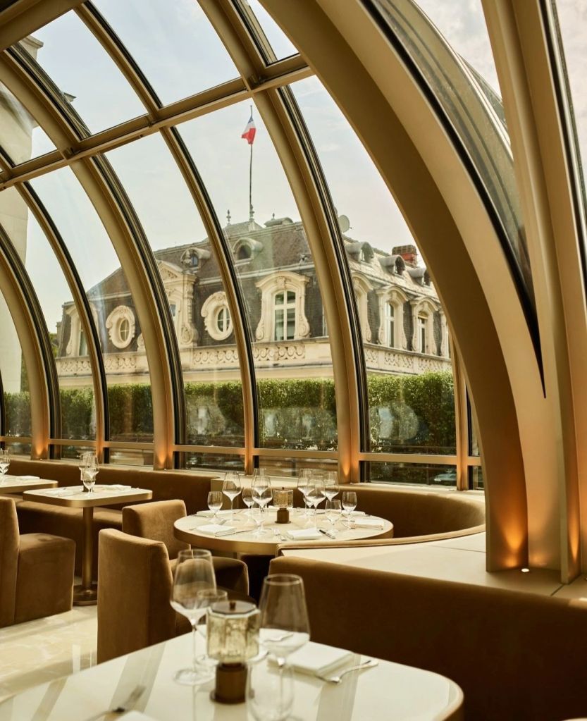 Interior view of a stylish restaurant with large arched windows offering a view of a Parisian building and flag, featuring elegantly set tables.
