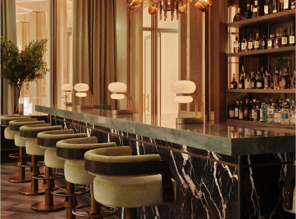A stylish bar area with green upholstered stools, marble countertop, modern lighting, and a shelf filled with various bottles.