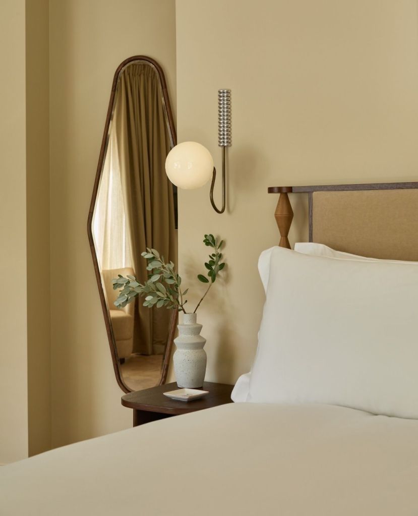 A cozy hotel bedroom showcasing a neutral color palette, featuring a comfortable bed, a decorative vase with foliage, a modern light fixture, and a decorative mirror reflecting soft light from a window.