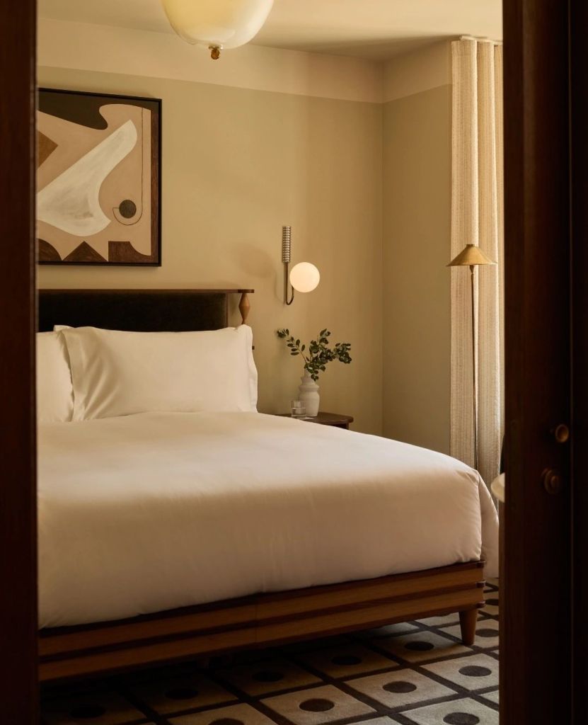 A warmly designed hotel bedroom featuring a large white bed, soft lighting, and abstract artwork on the wall.