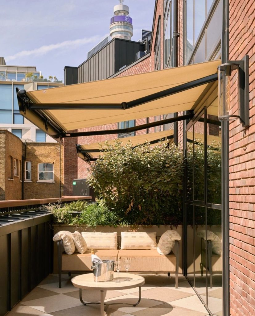 A stylish outdoor terrace at The Newman hotel featuring a comfortable seating area with cushions, a round table, and a bottle of champagne. Lush greenery surrounds the space, complemented by a modern building backdrop.