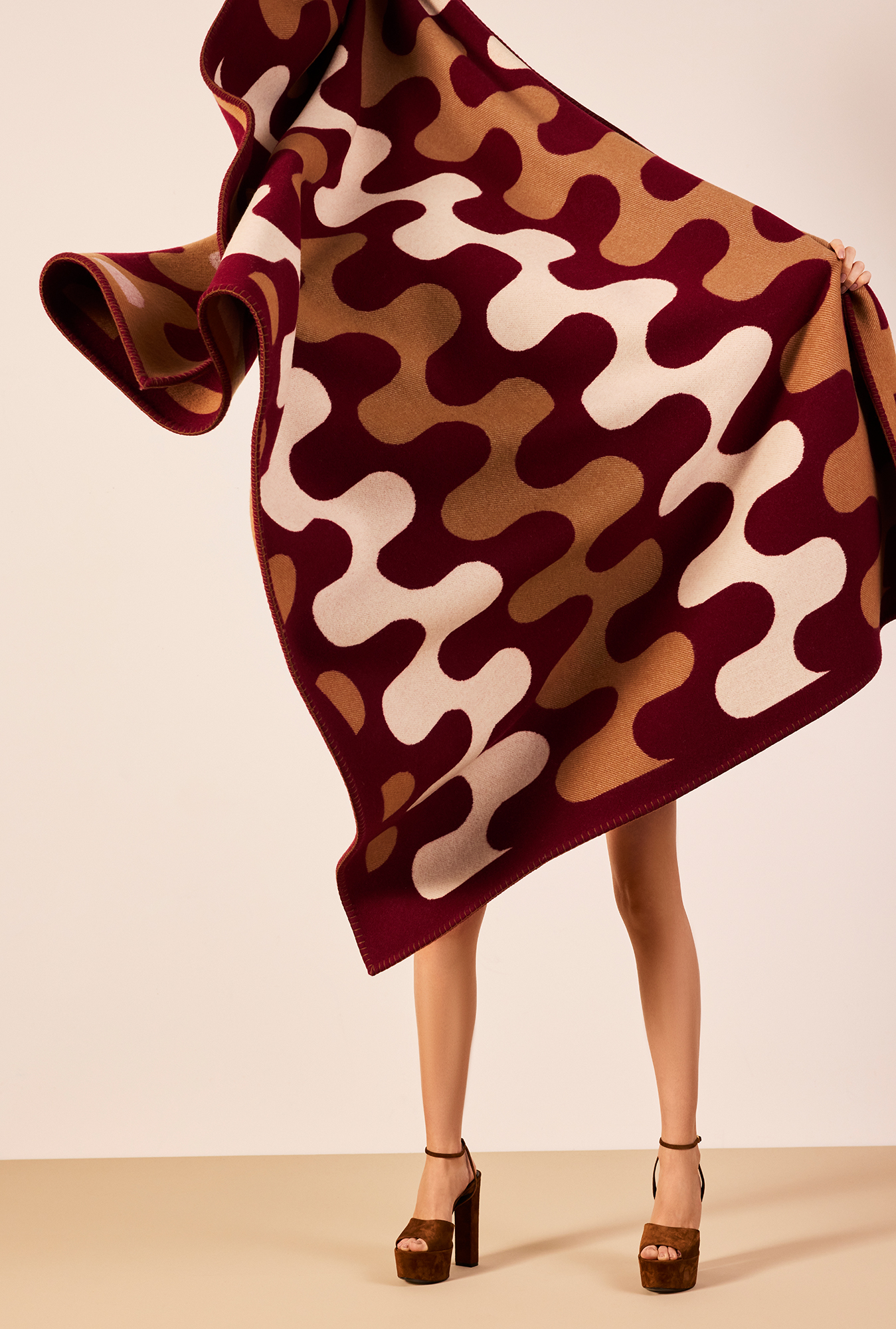 A jacquard-woven throw blanket featuring a wavy pattern in burgundy, tan, and cream colors, being held by a person in stylish platform shoes against a neutral background.