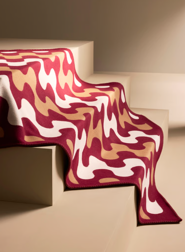 A jacquard-woven throw in warm tones of burgundy, tan, and white, draped over a set of neutral-colored stairs, showcasing a modern wave pattern.