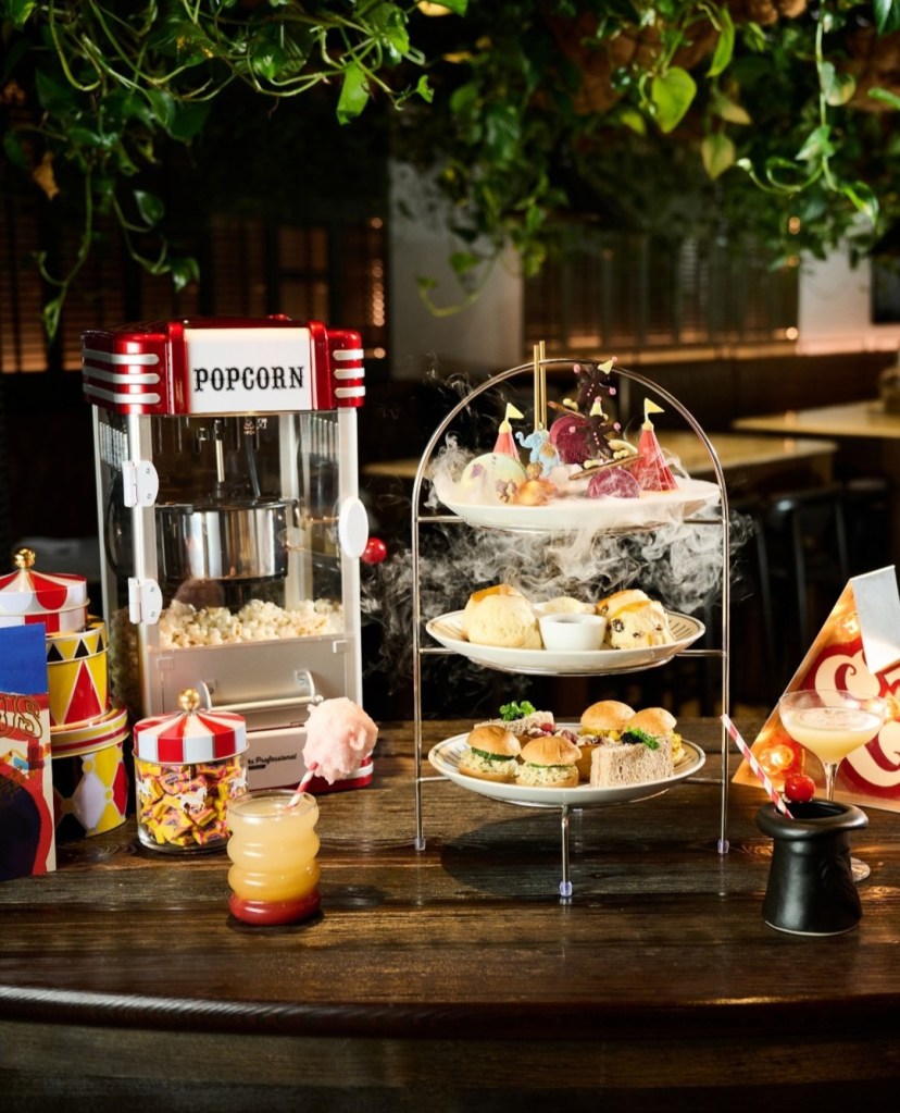 A creative afternoon tea display featuring a popcorn machine, colorful treats, scones, and savory sandwiches, all set on a wooden table decorated with playful party elements.