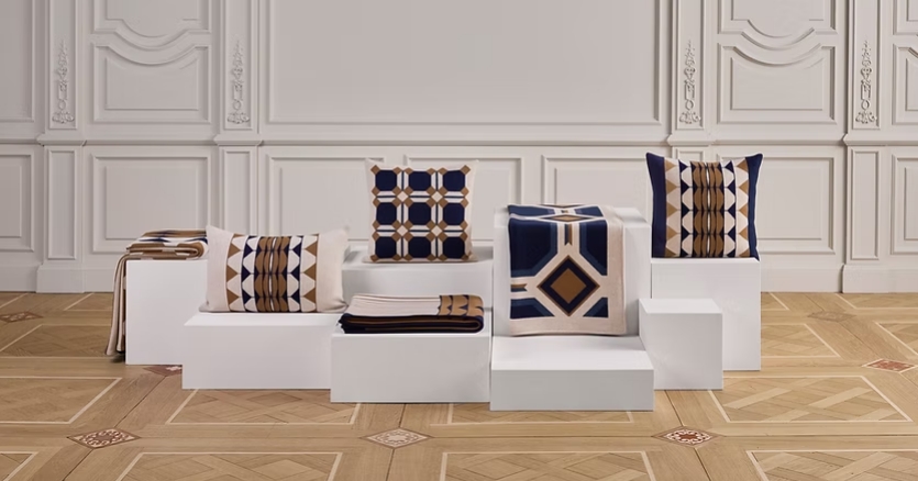 A collection of jacquard-woven throws and cushions arranged on white display platforms, featuring geometric patterns in shades of tan, navy blue, and cream, set against an elegant interior with ornate walls and wooden flooring.