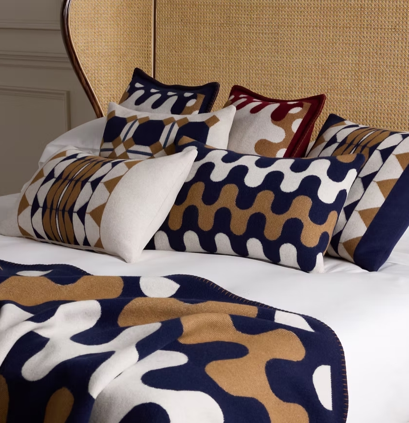 A close-up view of a beautifully arranged bed featuring various decorative cushions and a soft throw blanket, showcasing geometric and abstract patterns in rich colors such as navy blue, beige, and burgundy.