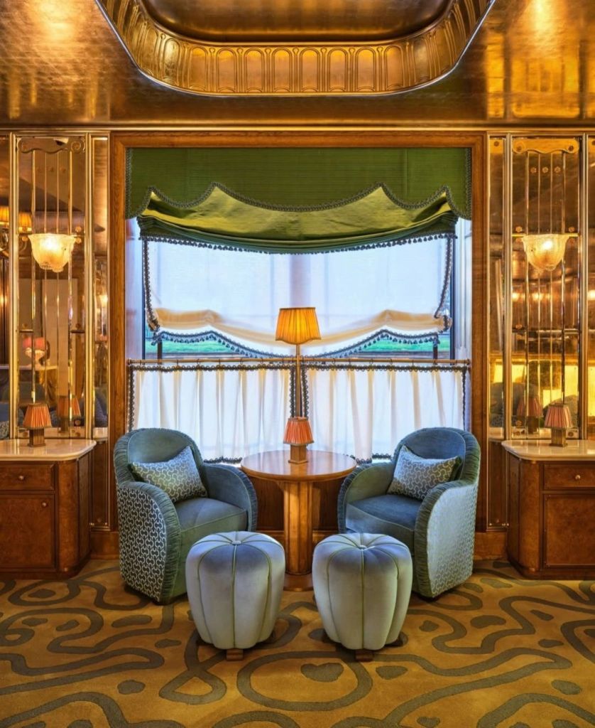 Elegant seating area in The Dorchester featuring plush blue armchairs, patterned cushions, and decorative light fixtures, with a round wooden table and stylish ottomans.