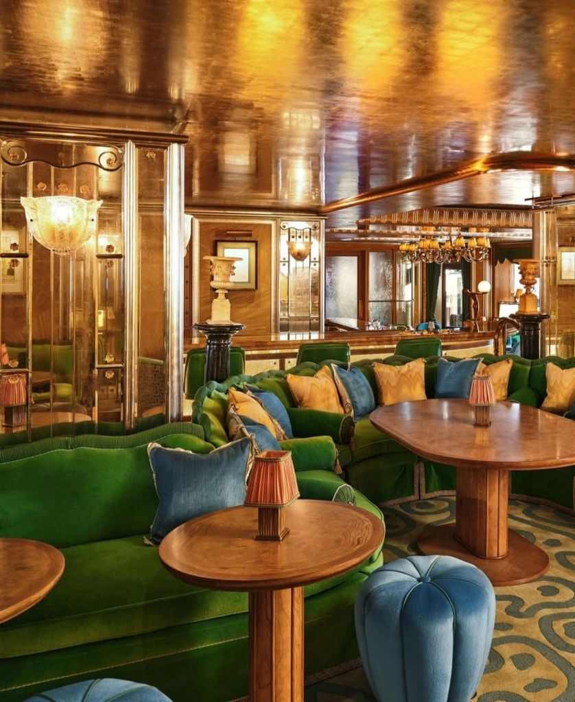 A luxurious lounge area featuring deep green curved sofas with colorful cushions, round wooden tables, and unique blue upholstered stools, all highlighted by warm golden lighting and elegant decor.