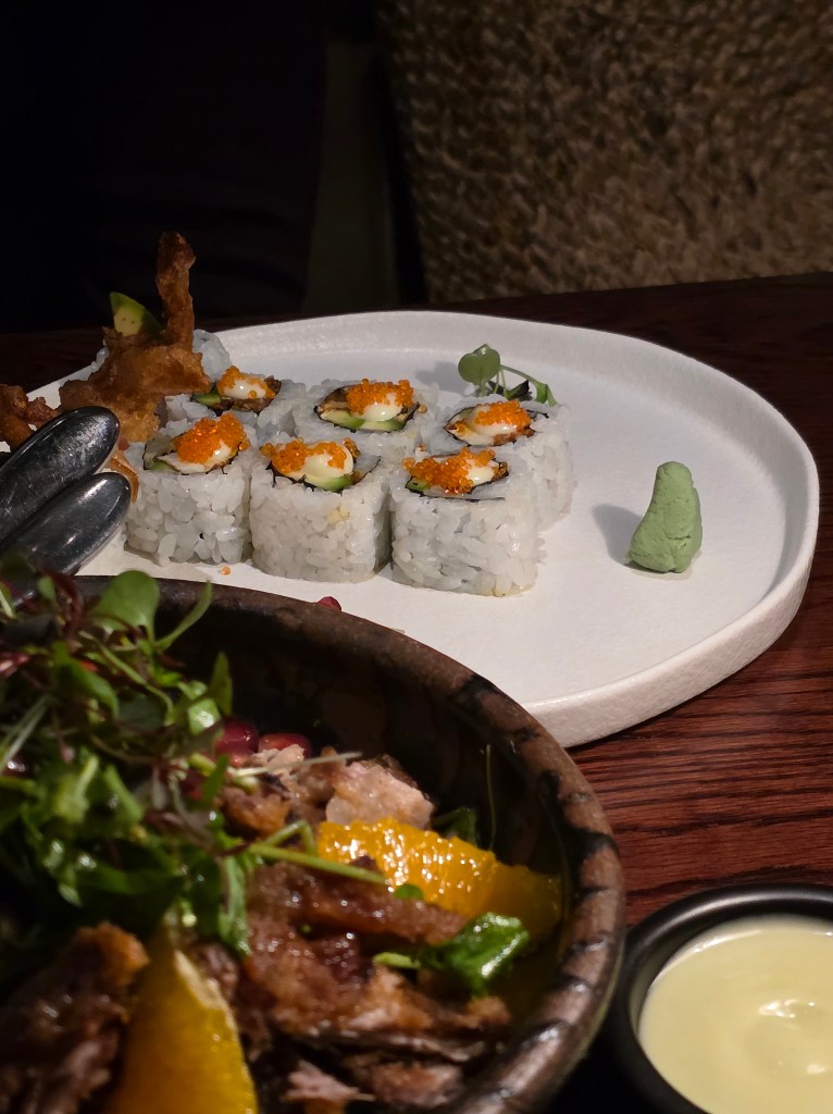 A white plate displaying sushi rolls topped with orange roe and accompanied by wasabi, with a green salad in a bowl in the foreground.