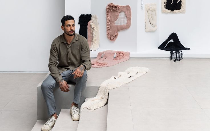A man sitting on a stair in a contemporary gallery, surrounded by various textile artworks displayed on the wall.
