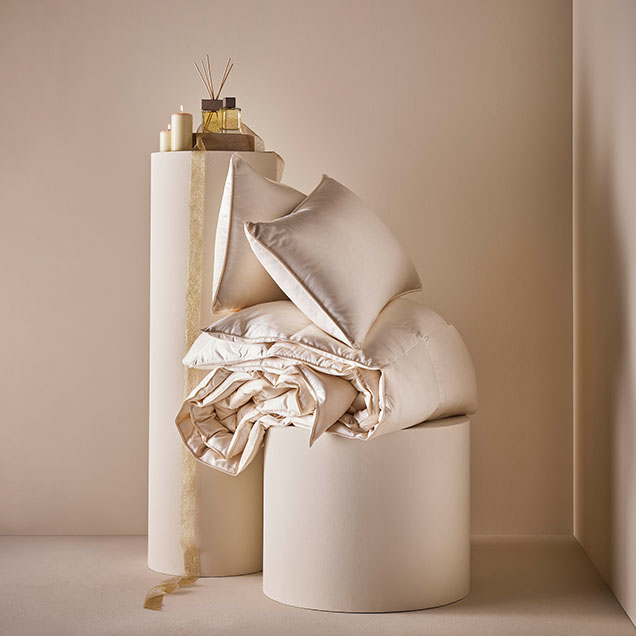 A serene display of luxurious bedding and cushions in soft cream tones, complemented by candles and decorative items on a minimalist pedestal.