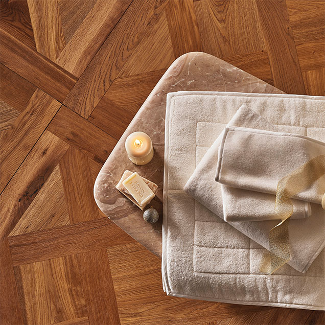 A flat lay arrangement featuring soft, neutral-toned towels and a delicate ribbon on a marble tray, accompanied by a lit candle and a bar of soap, set against a warm wooden floor.