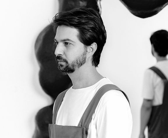 Black and white portrait of a man with medium-length hair and a beard, wearing a white shirt and overalls, standing against an abstract background.