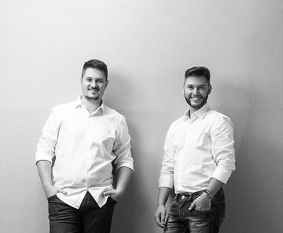 Two men standing side by side, both wearing white button-up shirts and jeans, with a plain light background.