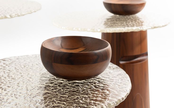 A wooden bowl sits atop a textured glass tabletop, emphasizing the contrast between natural and contemporary materials. Additional wooden elements are visible in the background.