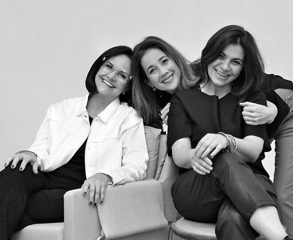 Three women posing together, smiling, in a monochrome setting. They are seated on a modern couch, dressed in stylish attire, exuding a friendly and relaxed atmosphere.