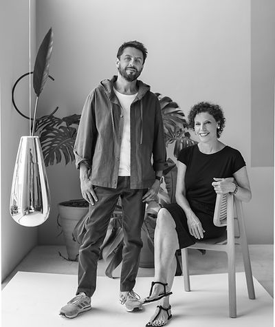 A black and white photograph of two individuals standing together in an indoor space decorated with plants and modern design elements, with one person seated on a chair.