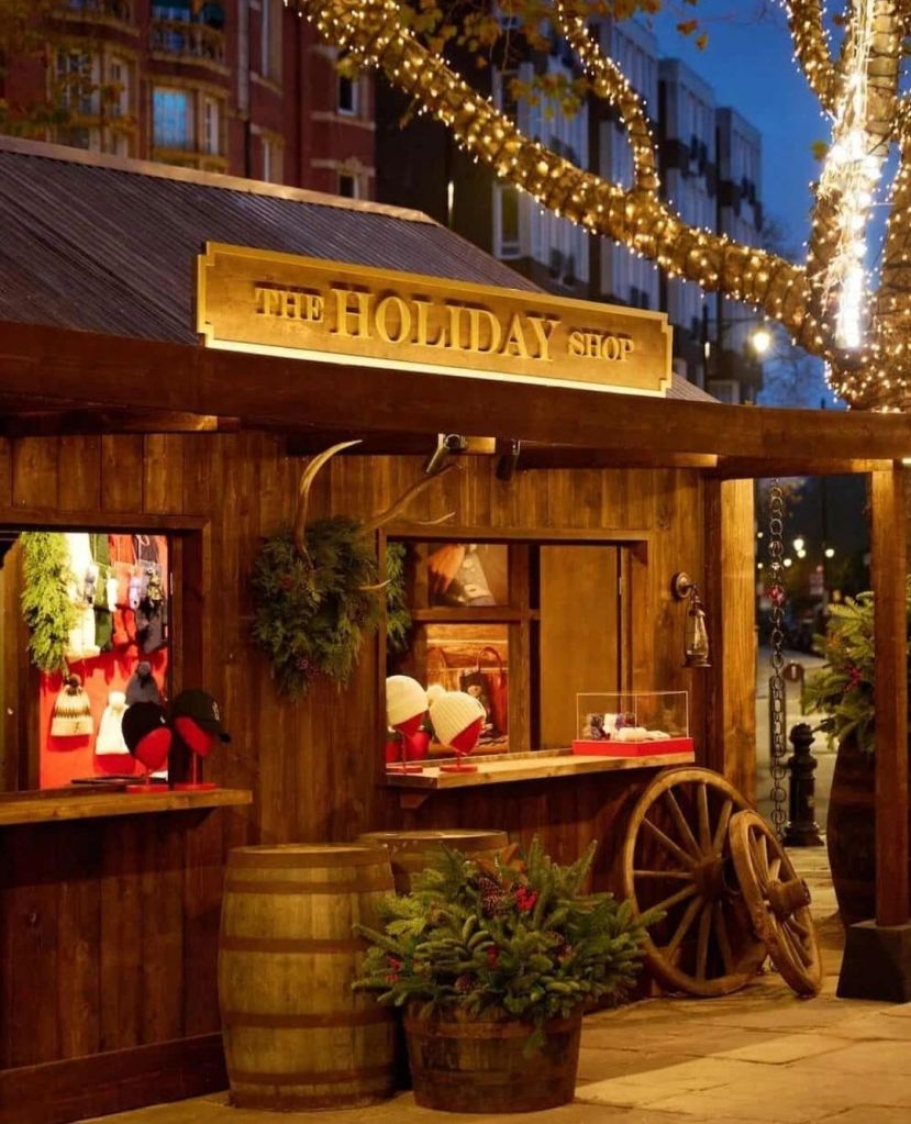 A festive wooden holiday shop adorned with string lights, featuring windows displaying winter accessories and wreaths, surrounded by rustic barrels and decorative plants.