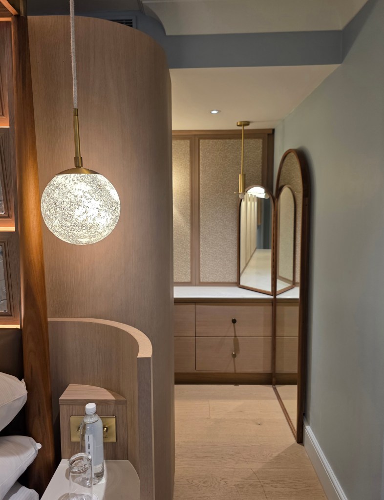 A cozy, elegantly designed hotel suite interior featuring a round pendant light, wooden accents, and a comfortable sleeping area, with an adjoining space showcasing a large mirror and minimalist cabinetry.