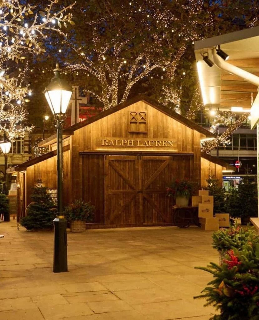 A beautifully lit rustic barn with 'RALPH LAUREN' displayed prominently, surrounded by winter pines and festive lights in Sloane Square, London.