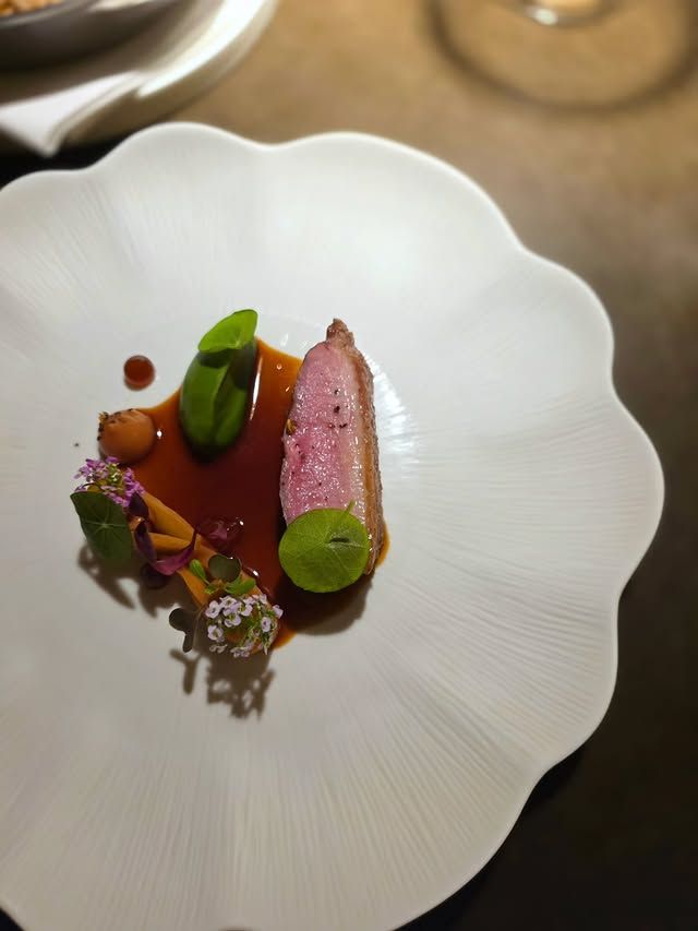 A beautifully plated dish featuring a slice of pink meat, garnished with fresh herbs and edible flowers, served in an elegant white bowl with sauce drizzled around.